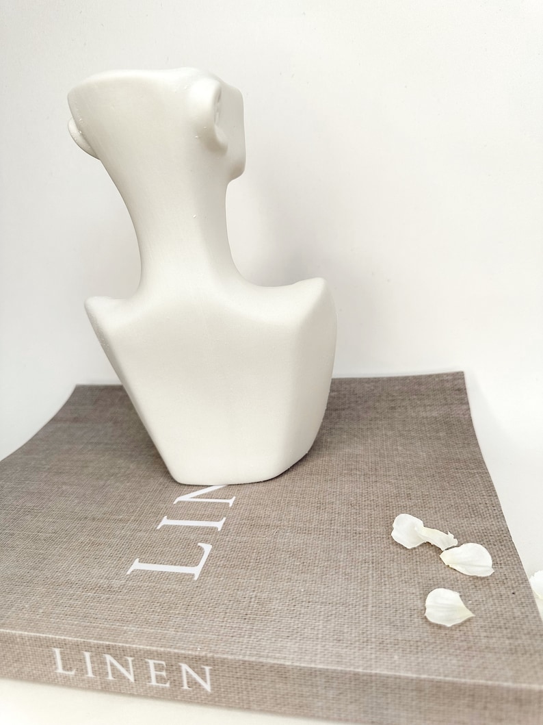 May include: White ceramic bust sculpture with a geometric design. The bust is sitting on a brown book with the word "LINEN" printed on the cover.