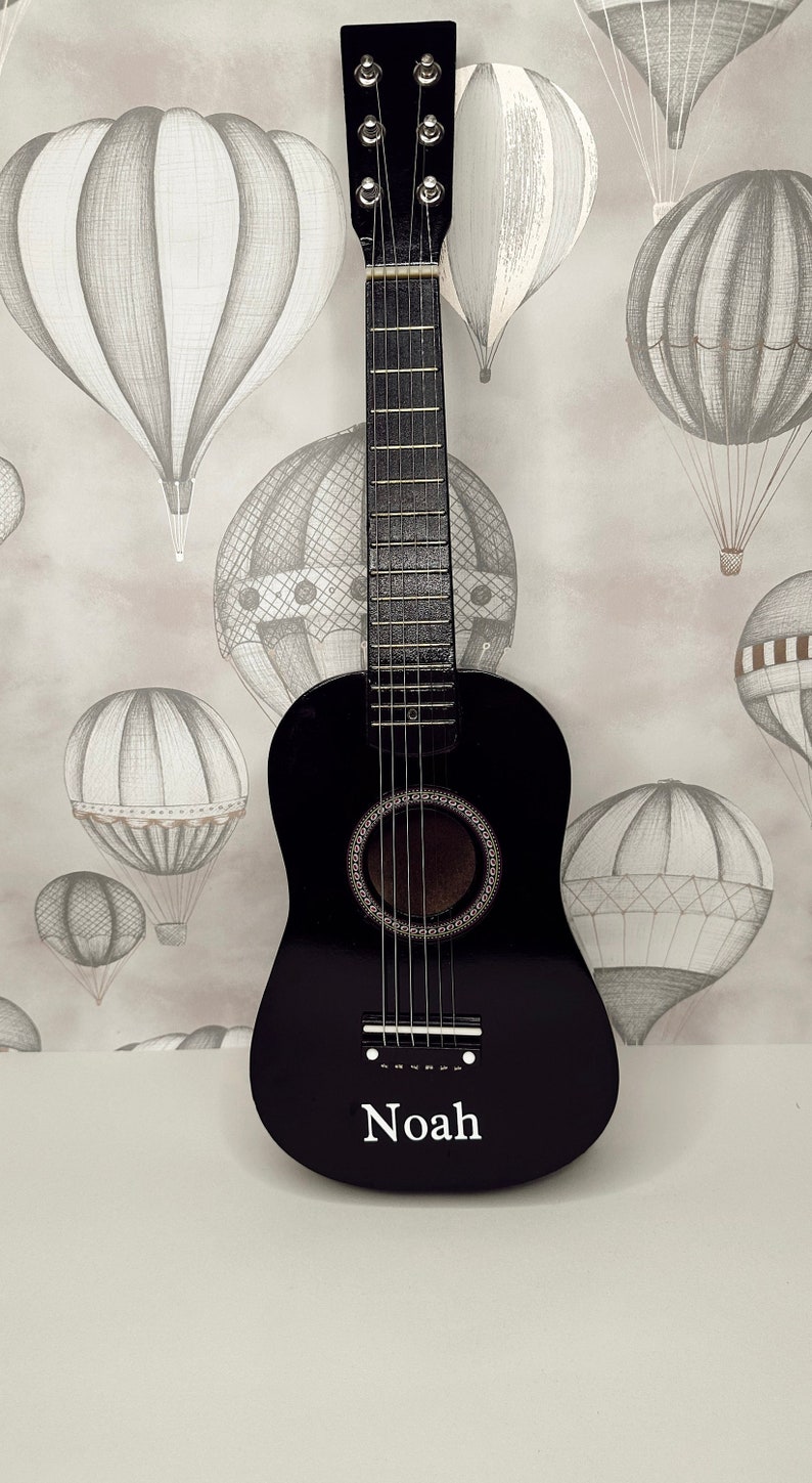 May include: A black acoustic guitar with a white name "Noah" on the front. The guitar is in front of a light gray background with a pattern of black and white hot air balloons.