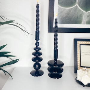 Può includere: Due portacandele neri con candele nere. I portacandele sono in vetro e hanno un design impilato unico. Le candele sono accese ed emanano una luce calda.