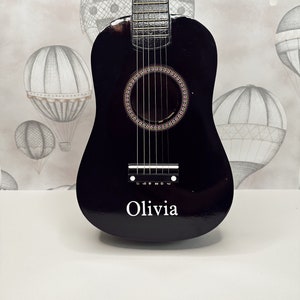 May include: A black acoustic guitar with a soundhole and six strings. The guitar has the name "Olivia" written on the front.