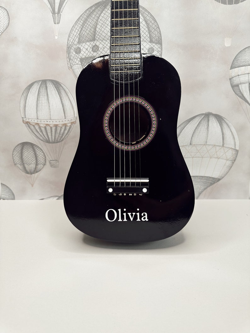 May include: A black acoustic guitar with a soundhole and six strings. The guitar has the name "Olivia" written on the front.