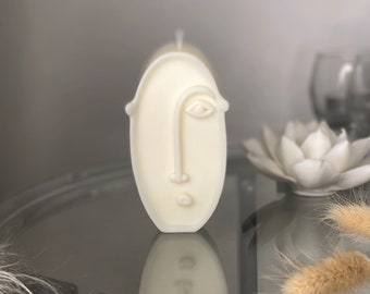 May include: White oval candle with a stylized face design. The face features a single eye, a nose, and a mouth.