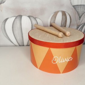 以下が含まれることがあります： 赤色のラインと「Olivia」という名前が白で書かれた、オレンジと黄色の小さなドラム。ドラムの上に木製のドラムスティックが2本置かれている。