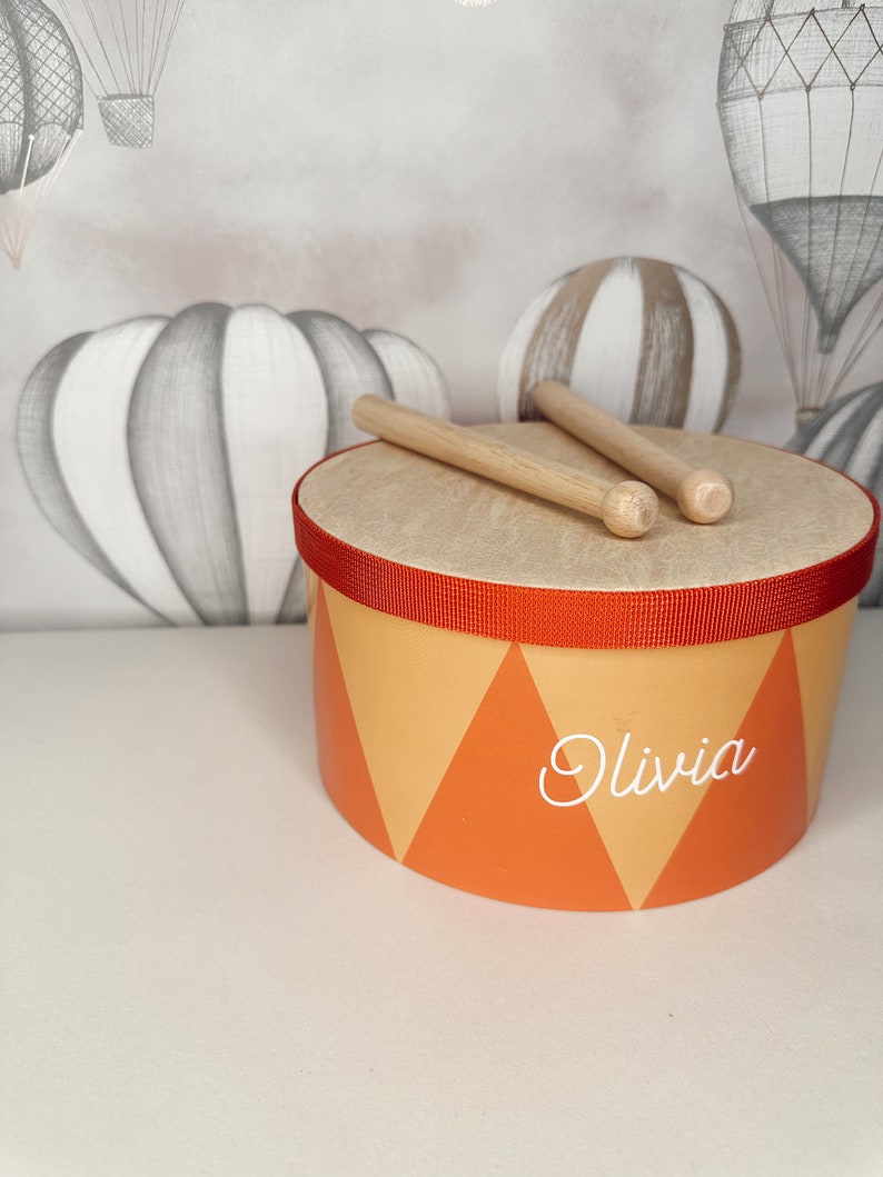 以下が含まれることがあります： 赤色のラインと「Olivia」という名前が白で書かれた、オレンジと黄色の小さなドラム。ドラムの上に木製のドラムスティックが2本置かれている。