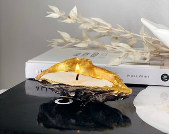 May include: A gold-painted oyster shell candle with a white wax interior and a lit wick. The candle is sitting on top of a black book with the word "CHANEL" printed on the front.