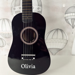 May include: A black acoustic guitar with a personalized inscription that reads "Olivia". The guitar has a light wood fretboard and a circular sound hole with a decorative inlay.