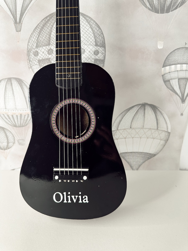 May include: A black acoustic guitar with a personalized inscription that reads "Olivia". The guitar has a light wood fretboard and a circular sound hole with a decorative inlay.