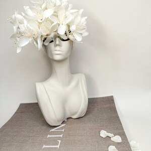 May include: White ceramic bust vase with a white flower arrangement on top. The vase is sitting on a book with the word "LINEN" printed on the cover.