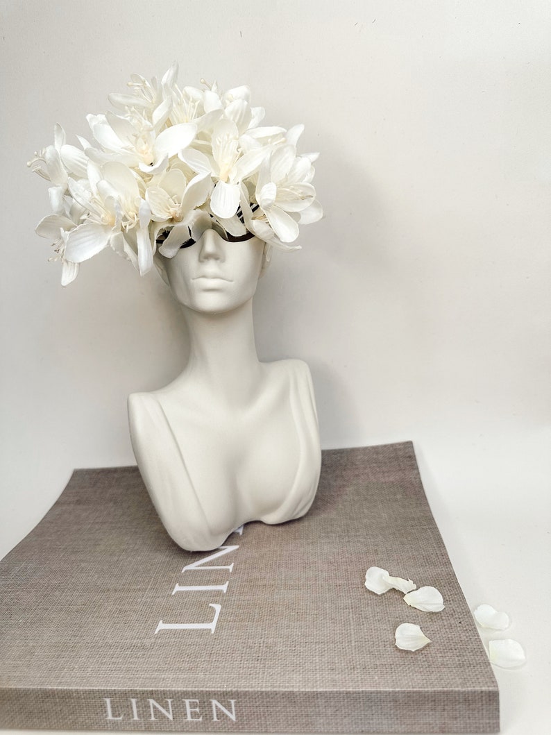May include: White ceramic bust vase with a white flower arrangement on top. The vase is sitting on a book with the word "LINEN" printed on the cover.