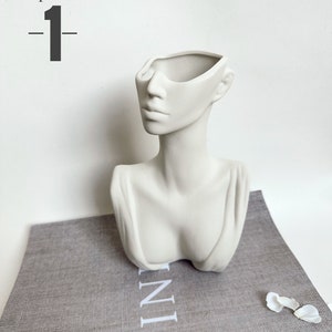 May include: White ceramic planter shaped like a bust of a woman with the top of the head removed to create a planter. The planter is sitting on a gray textured surface.