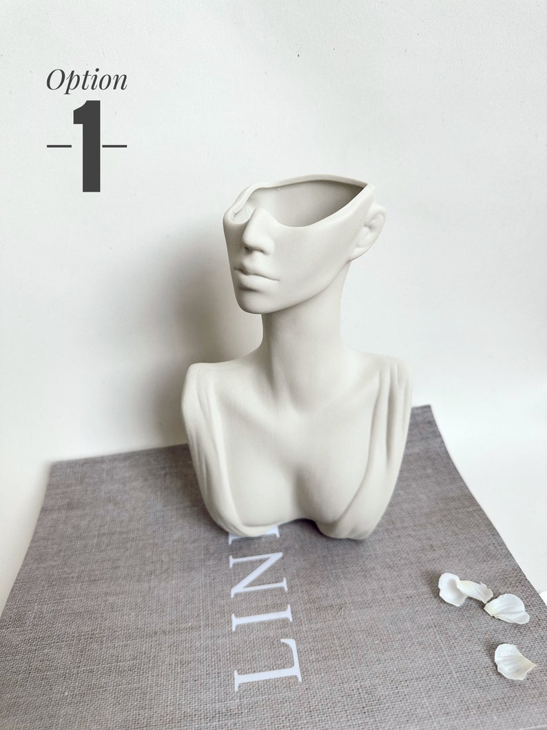 May include: White ceramic planter shaped like a bust of a woman with the top of the head removed to create a planter. The planter is sitting on a gray textured surface.