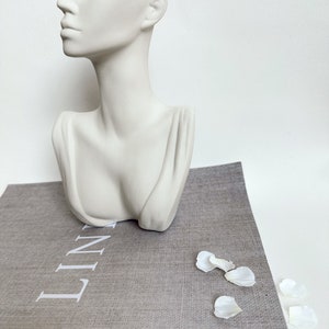 May include: White ceramic bust planter with a hollowed-out head, resembling a woman's face. The planter is sitting on a gray linen book with the word "LINEN" printed on the cover.