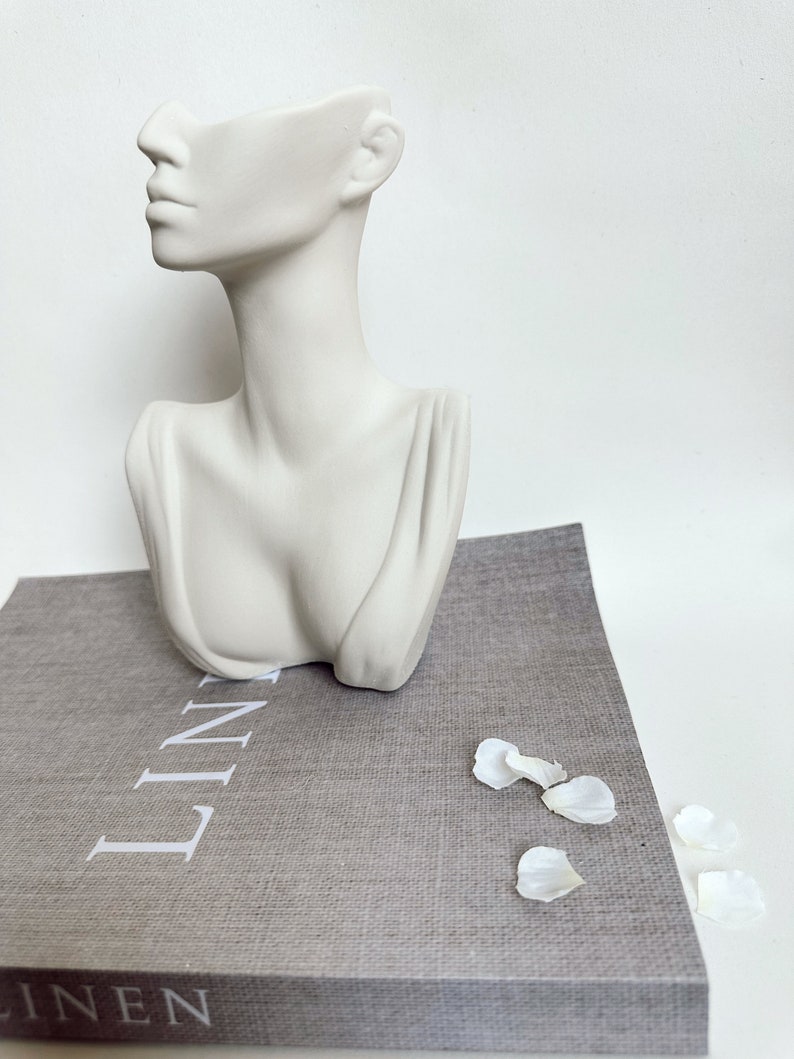 May include: White ceramic bust planter with a hollowed-out head, resembling a woman's face. The planter is sitting on a gray linen book with the word "LINEN" printed on the cover.