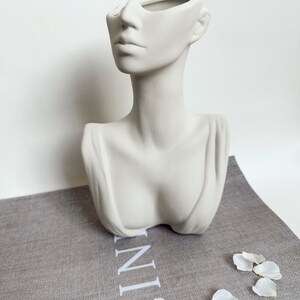 May include: White ceramic bust planter shaped like a woman's head with the top of the head removed to create a vase. The planter is sitting on a gray linen surface with white flower petals scattered around it.