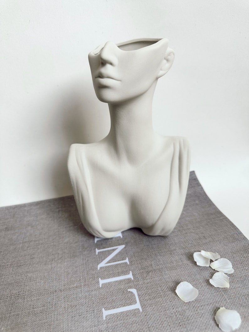 May include: White ceramic bust planter shaped like a woman's head with the top of the head removed to create a vase. The planter is sitting on a gray linen surface with white flower petals scattered around it.