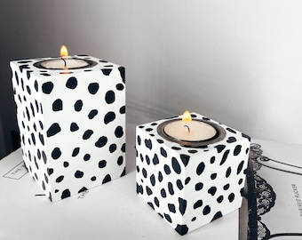 May include: Two white candle holders with a black and white dalmatian print design. The candle holders are square and have a small opening in the center for a tea light candle. The candle holders are sitting on a white book with the word "WHITE" printed on the cover.