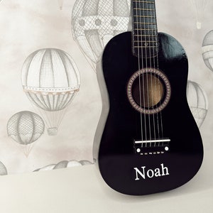 May include: A black acoustic guitar with a name "Noah" inscribed on the front. The guitar is in front of a light pink and white background with a pattern of hot air balloons.