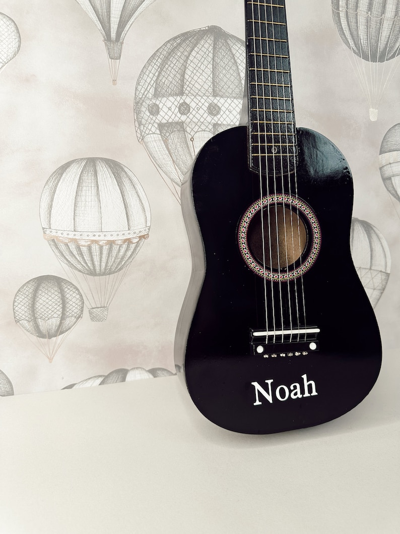 May include: A black acoustic guitar with a name "Noah" inscribed on the front. The guitar is in front of a light pink and white background with a pattern of hot air balloons.