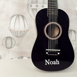 May include: A black acoustic guitar with a white inscription that reads "Noah" on the body. The guitar is in front of a wall with a light brown background and a pattern of gray hot air balloons.