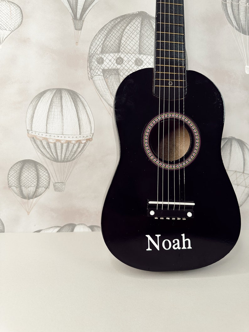 May include: A black acoustic guitar with a white inscription that reads "Noah" on the body. The guitar is in front of a wall with a light brown background and a pattern of gray hot air balloons.