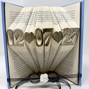 May include: A folded book art sculpture featuring the date 02-07-27 with heart shapes between the numbers. The pages are precisely creased to form the digits and hearts. A black ribbon with a small white crocheted flower is tied around the base.