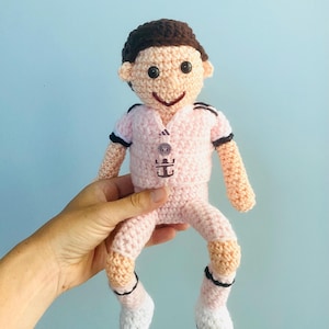 May include: A handmade crochet doll in a light pink outfit with black and white accents. The doll has a smiling face, brown hair, and is wearing a shirt and pants. The doll is being held against a light blue background.