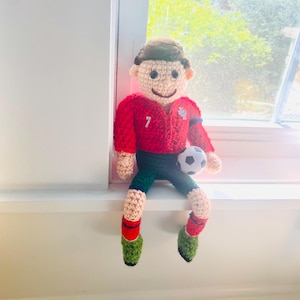 Puede incluir: Muñeco de ganchillo hecho a mano de un jugador de fútbol con camiseta roja con el número 7, pantalones cortos verdes y calcetines de fútbol. El muñeco sostiene un pequeño balón de fútbol. Hay otro balón debajo.