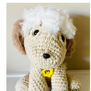 May include: A crocheted cream-colored dog with a white fluffy head, brown ears, and black eyes. The dog is wearing a yellow collar with a letter "M" on it.