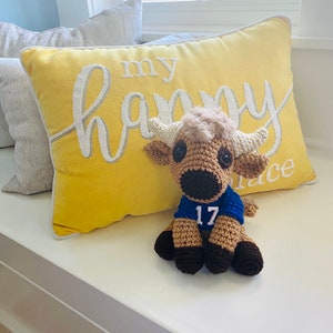 May include: A crocheted stuffed animal wearing a blue jersey with the number 17 on it. The stuffed animal is sitting on a yellow pillow with the text "my happy place" embroidered on it.