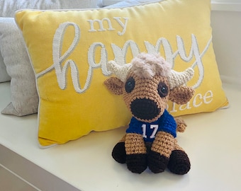 May include: A crocheted stuffed animal wearing a blue jersey with the number 17 on it. The stuffed animal is sitting on a yellow pillow with the text "my happy place" embroidered on it.