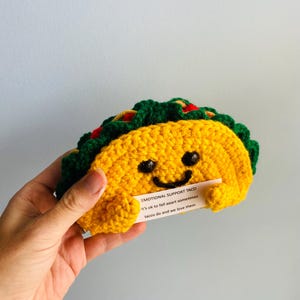 以下が含まれることがあります： 黄色、緑、赤色の手編みタコス。タコスは笑顔で、「EMOTIONAL SUPPORT TACO」と応援メッセージが書かれた小さなカードを持っています。楽しくてユニークなギフトです。