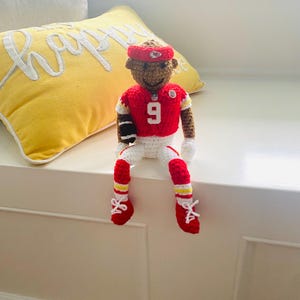 May include: A crocheted doll dressed in a red and white football uniform with the number 9 on the chest, a red headband with a logo, and red shoes with white laces. The doll has brown skin, holds a small brown football, and wears white gloves.