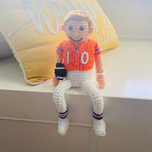 Op de afbeelding: Handgemaakte gehaakte pop van een American footballspeler met een oranje shirt met nummer 10. De pop heeft een witte broek, blauw-oranje gestreepte sokken en houdt een zwart-witte football vast. De pop zit op een wit oppervlak.