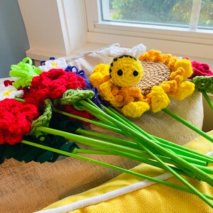 May include: A collection of handmade crochet flowers in various colors, including red, white, and yellow. A yellow crochet bee sits atop a sunflower. The flowers have green stems and are arranged on a yellow cushion.