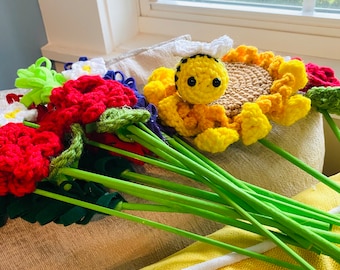 May include: A collection of handmade crochet flowers in various colors, including red, white, and yellow. A yellow crochet bee sits atop a sunflower. The flowers have green stems and are arranged on a yellow cushion.