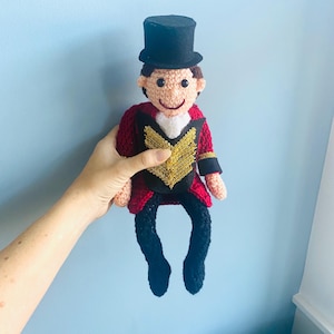 May include: Crocheted doll of a ringmaster wearing a black top hat, red jacket with gold accents, black pants, and a white shirt. The doll has a smiling face and is handmade. The doll is a circus-themed item.