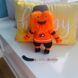 May include: A handmade orange and black crocheted mascot doll with a hockey stick. The doll has a black hat, orange fur, and a Flyers jersey. The hockey stick is white with red stripes and a black blade.