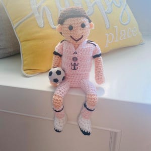 May include: A handmade crochet doll featuring a person with brown hair wearing a light pink soccer jersey with black trim, matching shorts, and white socks with black accents. The doll holds a small black and white soccer ball in its right hand.