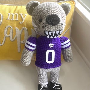 May include: A gray crocheted stuffed animal wearing a purple jersey with a white number "0" and a white K-State logo. The animal has a black nose and white teeth.