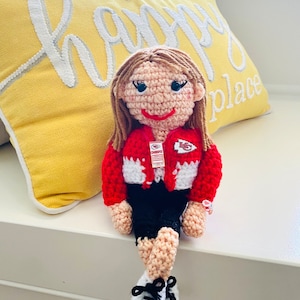 May include: A handmade crochet doll with light brown hair, wearing a red and white jacket, black shorts, and white sneakers with black laces. The doll has a smiling face and is sitting on a white surface. A yellow pillow with the words "my happy place" is in the background.