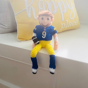 May include: A handmade crochet doll of a football player wearing a blue jersey with the number 9, yellow trousers, and a black and white football. The doll is sitting on a white surface with a yellow pillow in the background.