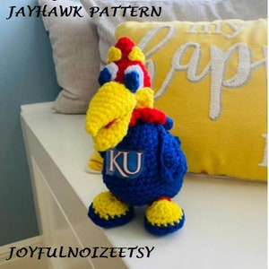 May include: Crocheted stuffed toy of the University of Kansas mascot, a blue jay, with a yellow beak, red and white crest, and a blue body with the letters "KU" on the chest. The toy is sitting on a white surface.