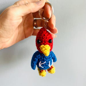 May include: A crocheted bird keychain with a red head, blue body, and yellow beak and feet. The bird is holding a small white heart in its feet.