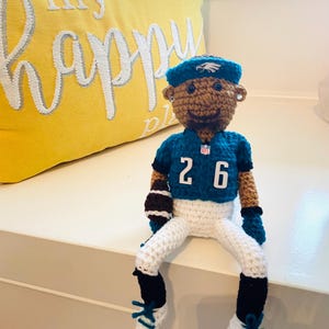 May include: Crocheted football player doll wearing a green and white jersey with the number 26 on the front. The doll is sitting on a white surface.