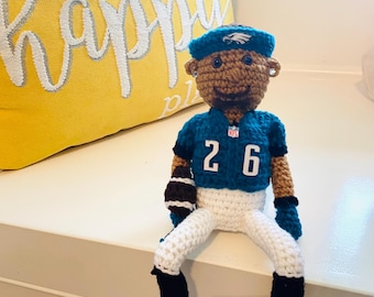 May include: Crocheted football player doll wearing a green and white jersey with the number 26 on the front. The doll is sitting on a white surface.