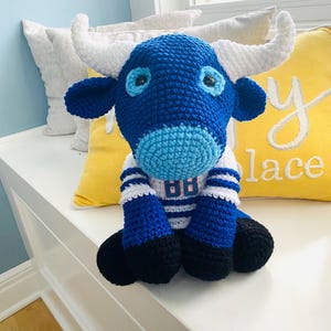 Op de afbeelding: Een handgemaakt gehaakt stierenspeelgoed in blauw, wit en zwart. De stier heeft grote witte hoorns, blauwe ogen en draagt een trui met het nummer "88". Het speelgoed staat op een wit oppervlak, met gele en witte kussens op de achtergrond.