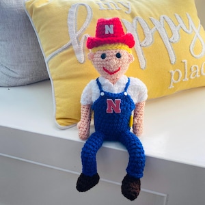 May include: A crocheted doll wearing a red hat with a white "N" on it, blue overalls with a red "N" on the front, and brown shoes. The doll is sitting on a yellow pillow with the words "my happy place" embroidered on it.