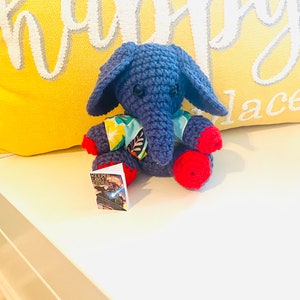 May include: A blue crocheted elephant wearing a colorful floral shirt and red crocheted shoes. The elephant is sitting on a white surface with a book titled "Keeper of Lost Cities" in the foreground.