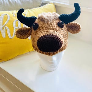 May include: A brown crocheted cow hat with black horns and brown eyes. The hat is on a white mannequin head.