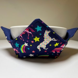 May include: A navy blue bowl cozy with a white ceramic bowl inside. The cozy features a colorful unicorn and rainbow pattern. The cozy has a square shape with flaps on each side to hold the bowl.